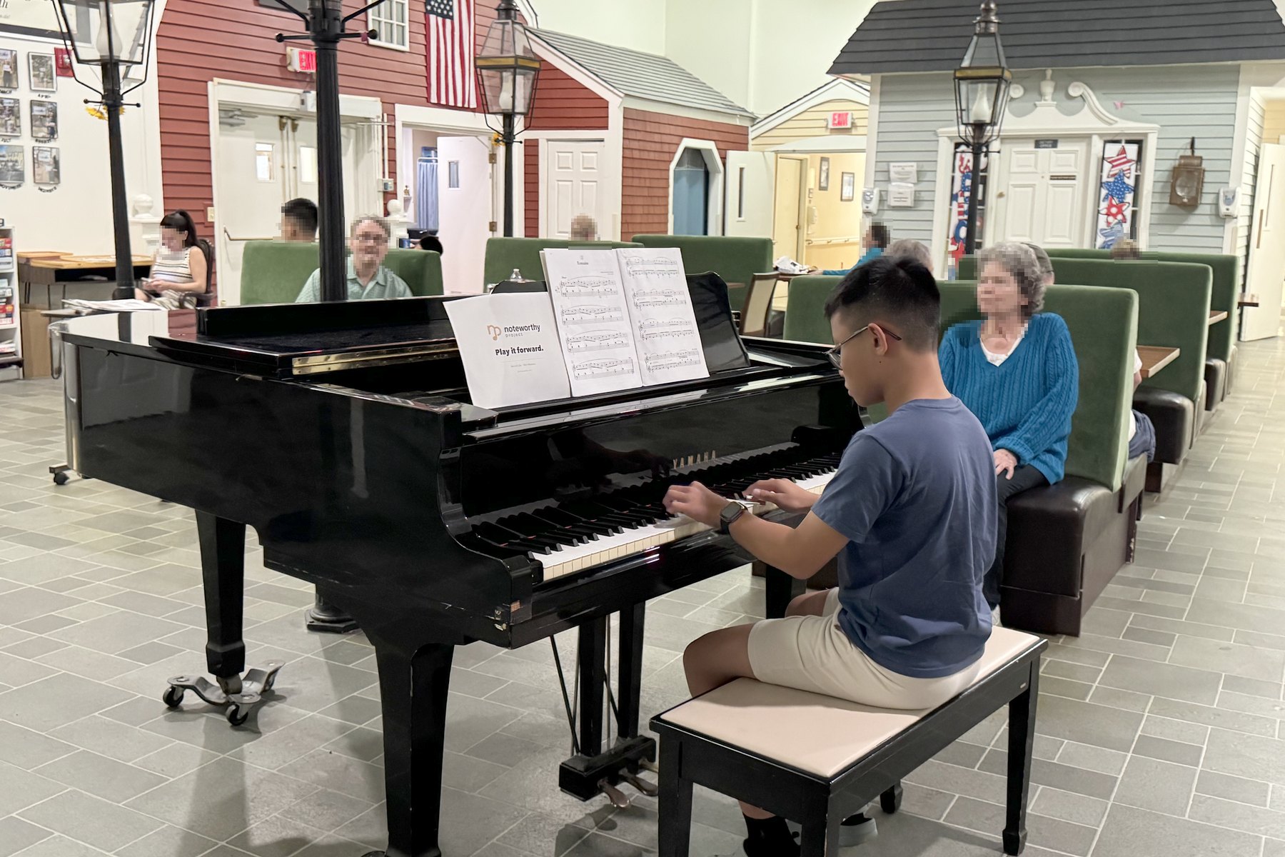 A student playing a grand piano in a residence common room, with people listening from the seating.
