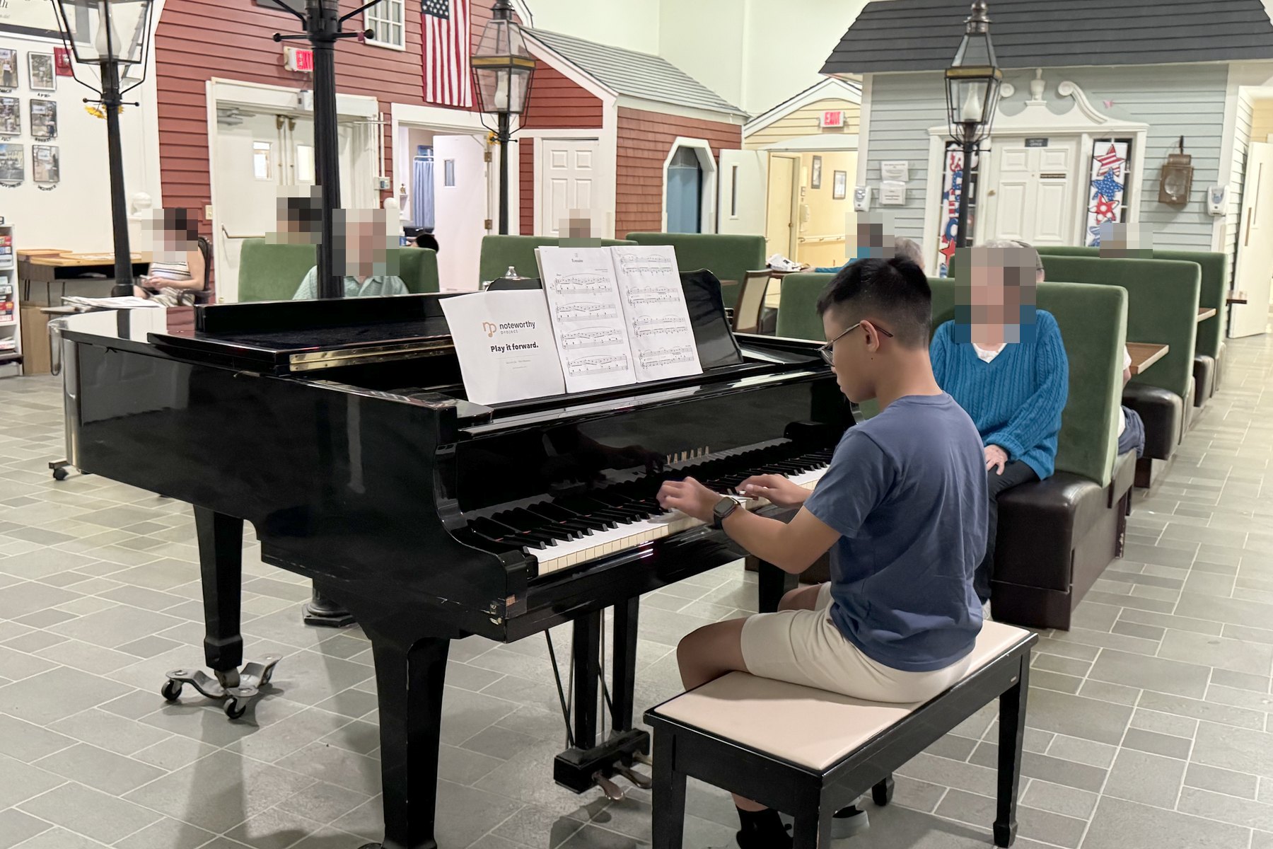 A student playing a grand piano in a residence common room, with people listening from the seating.