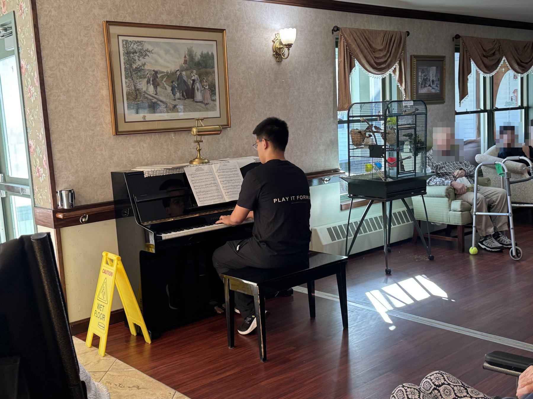 A student playing an upright piano in a senior community day room, with residents listening by the window.