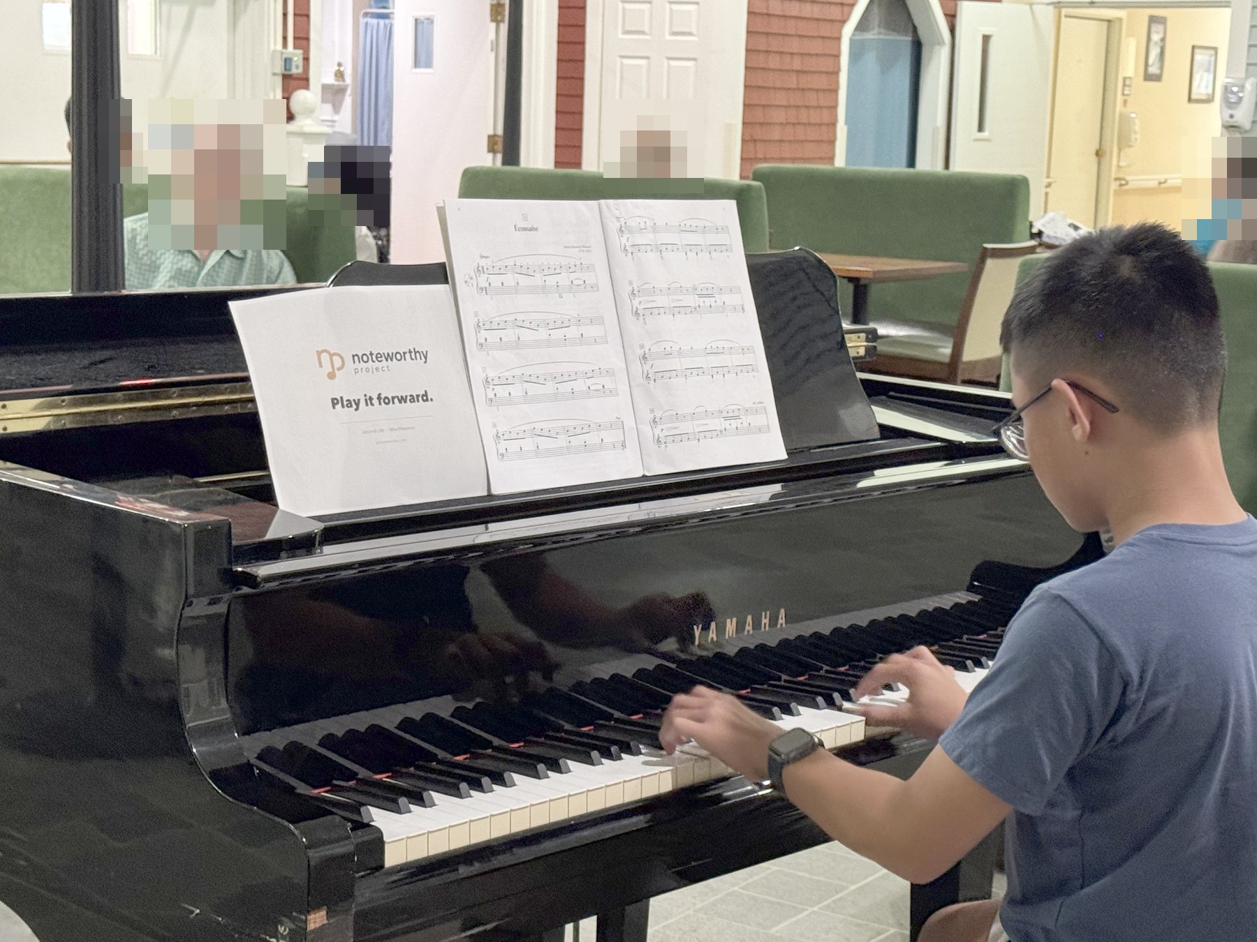 A student playing a grand piano from sheet music, in a residence day room.